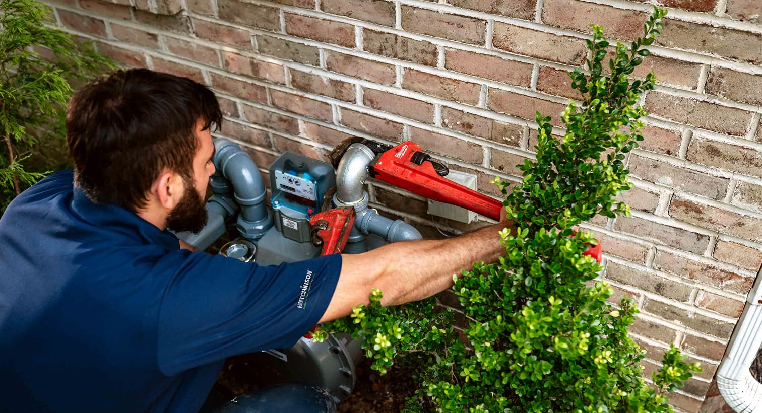 A professional Hutchinson technician using a red pipe wrench to service outdoor residential gas line plumbing and pipe connections against a brick wall.