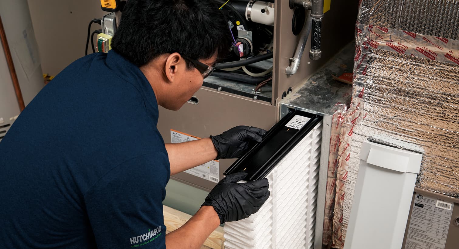 A professional Hutchinson HVAC technician replacing a clean pleated air filter in an indoor air handler unit during a routine AC maintenance tune-up.